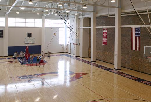 Basketball court with pine wood floors and exposed steel ceiling. Navy and red school logo on the floor.