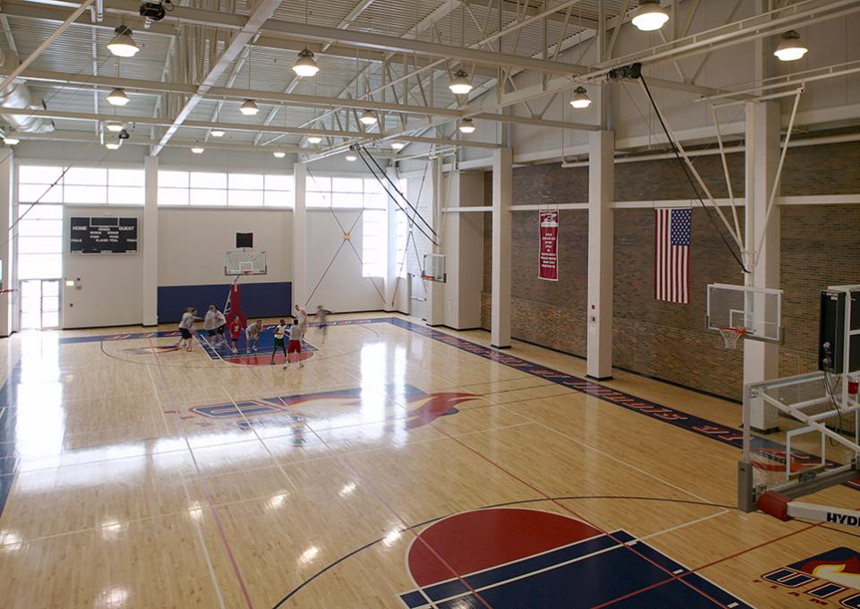 Basketball court with pine wood floors and exposed steel ceiling. Navy and red school logo on the floor.